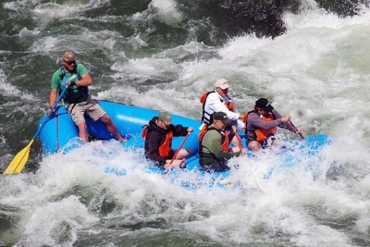 Group of six people white-water rafting in a blue raft on a turbulent river.